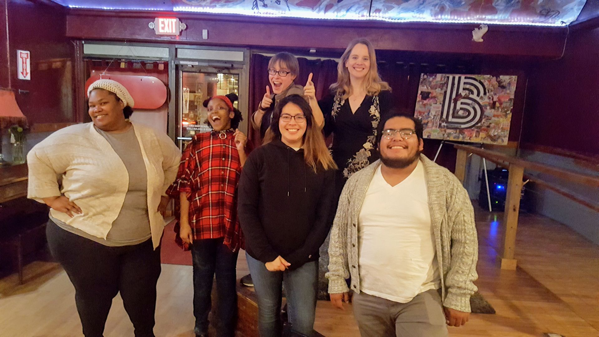 Five people smiling in a dim indoor venue with wood floors and a neon sign.
