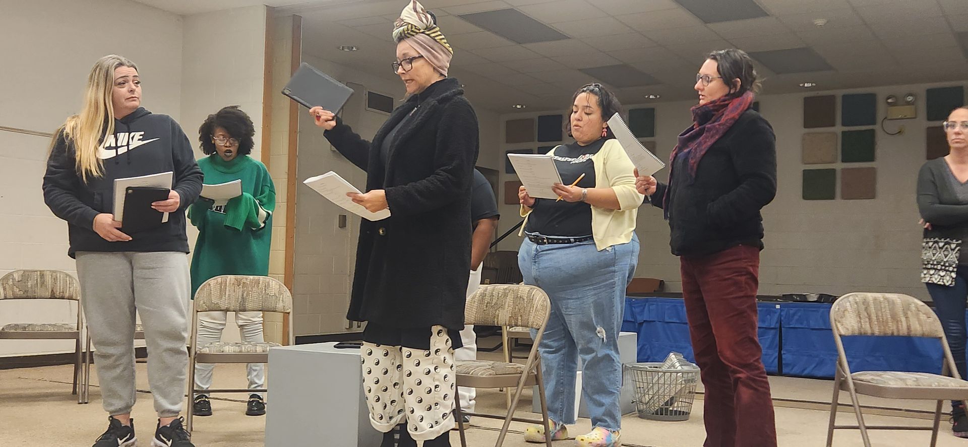 Five women stand on a stage reading papers during a group event in a community room.