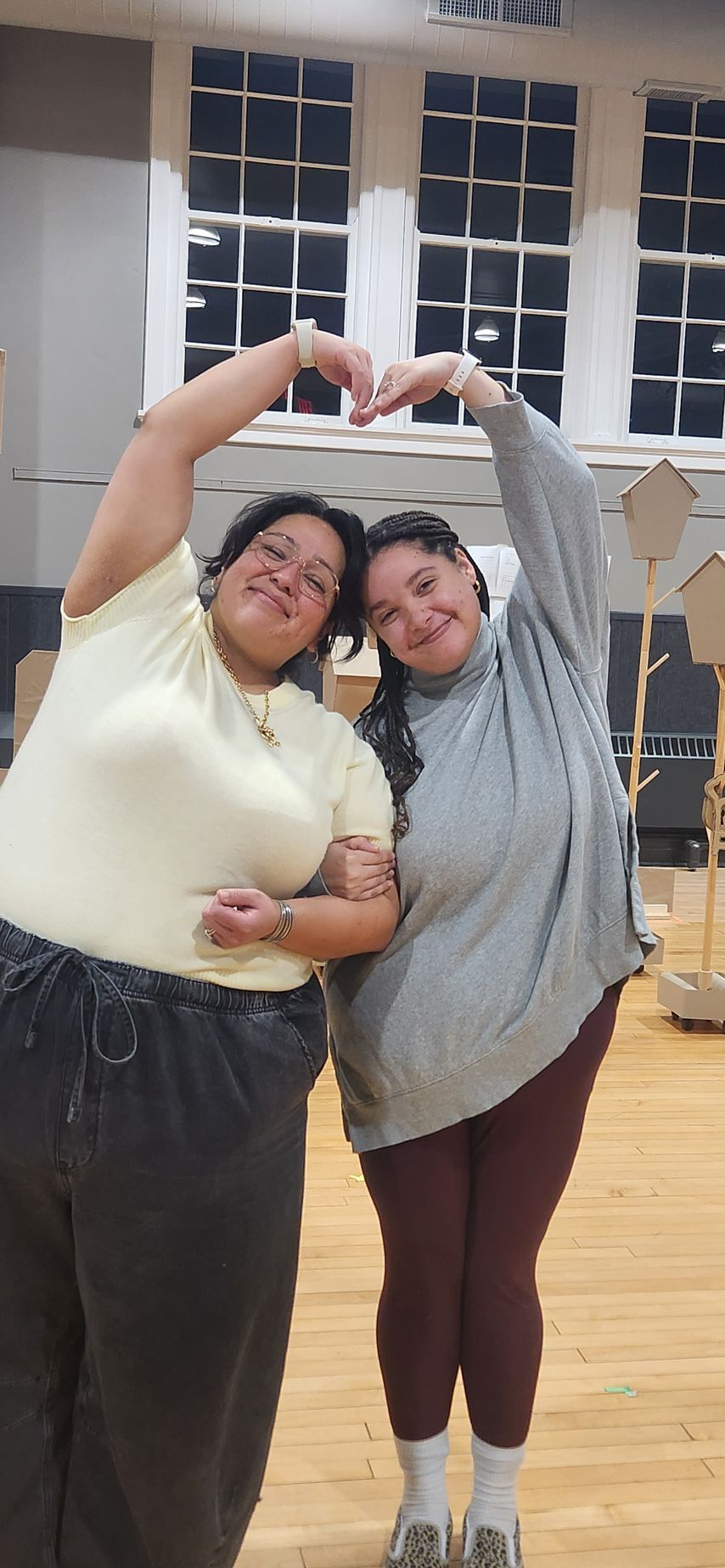 Two people in casual clothes stand indoors, smiling and forming a heart shape with their arms.