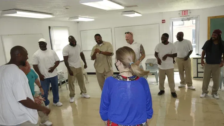 People standing in a circle in a bright room, appearing to participate in a group activity.