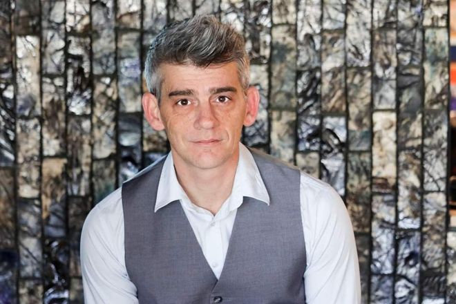 Person in a white shirt and gray vest seated against a patterned curtain background.