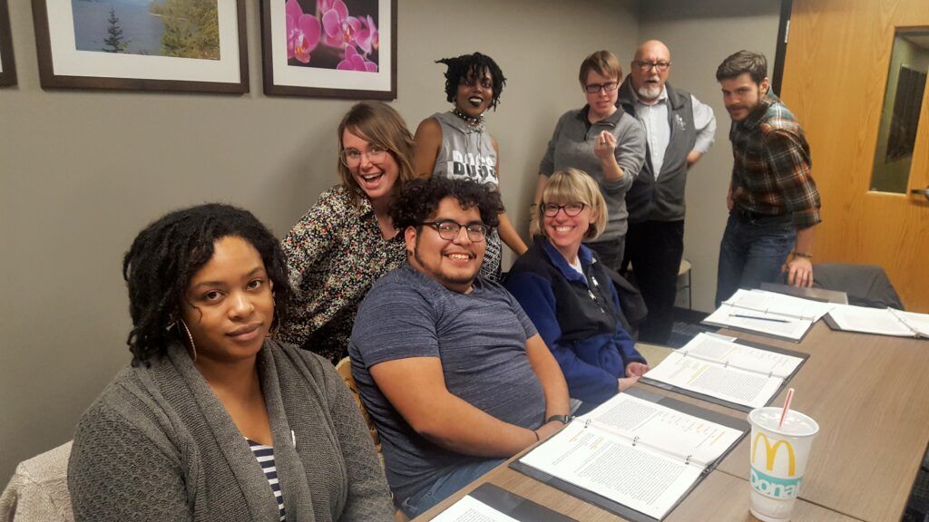People seated at a conference table in an office, with documents and a cup in front of them, smiling at the camera.