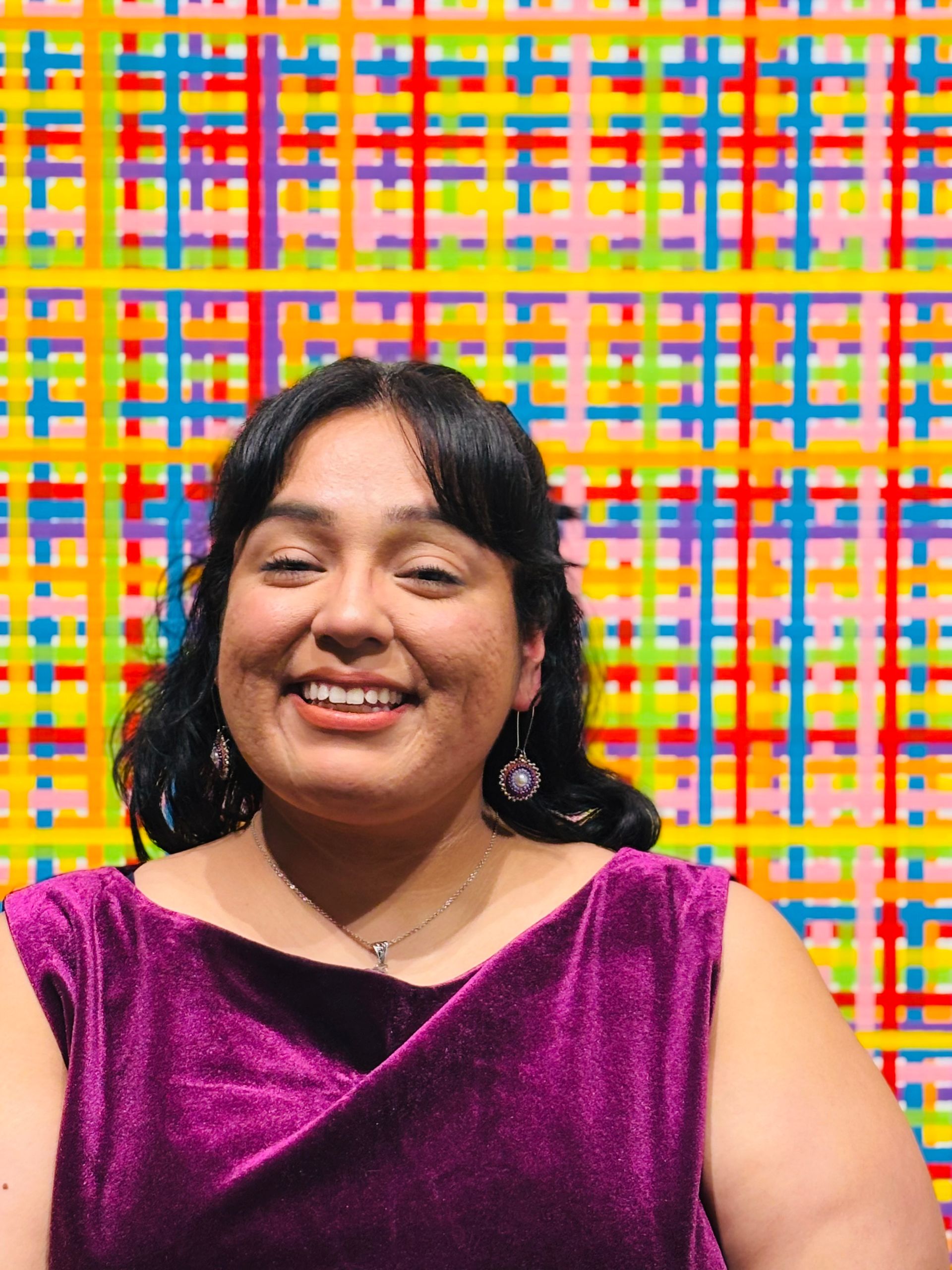 Smiling person in a purple velvet top in front of a colorful woven-pattern backdrop