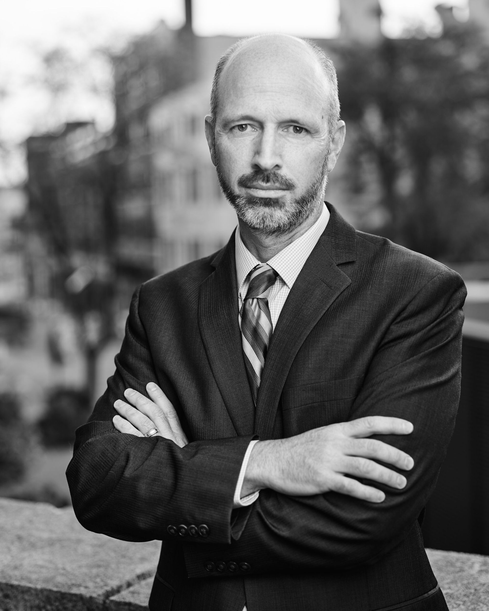 Portrait of a bald man in a dark suit and tie, standing with arms crossed outdoors.