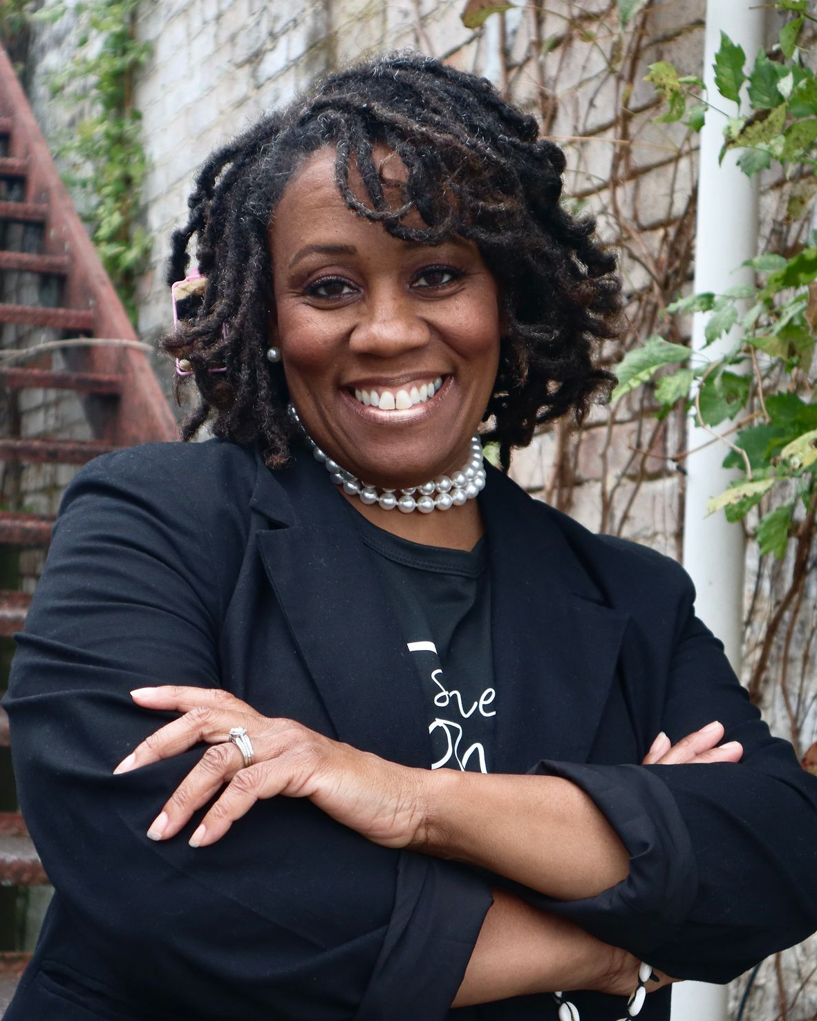 Smiling person in black blazer with arms crossed, standing beside outdoor stairs and ivy-covered wall.