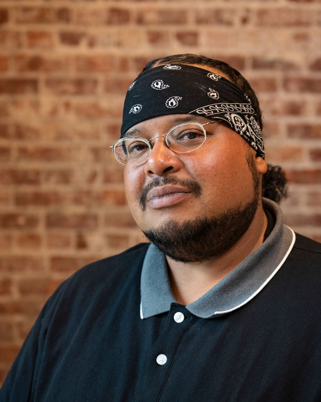 Man in glasses and black bandana wearing a black polo, posed against a brick wall.