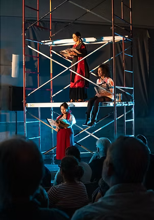 Three performers on scaffolding platform, one playing violin, under stage lights.