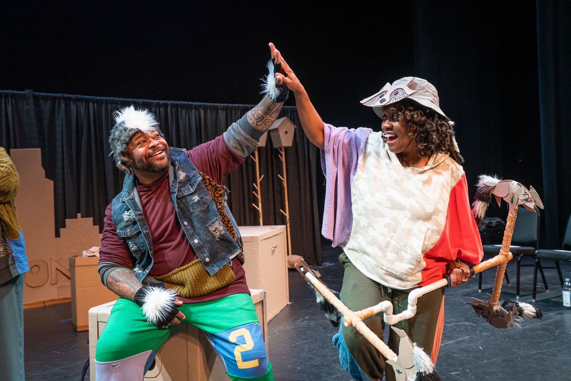 Two costumed performers high-five onstage, one holding a bicycle prop and the other a hatchet.