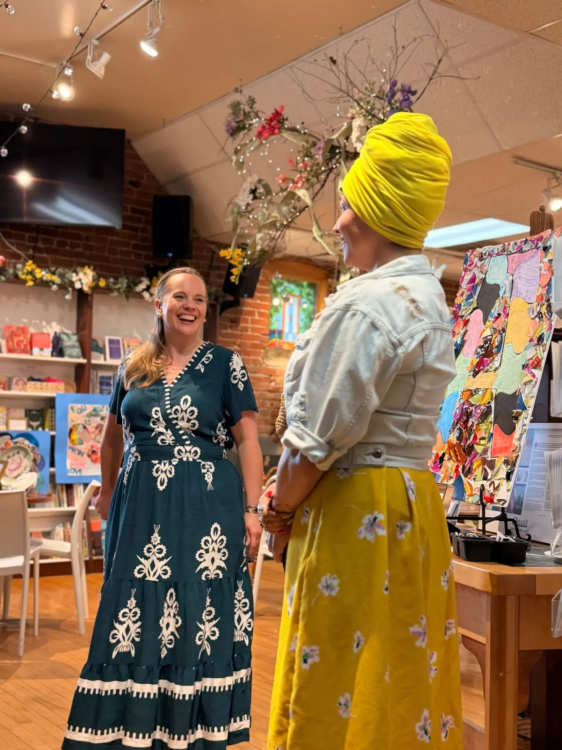 Two women smiling in a decorated shop, one in a teal dress and one in a yellow headwrap and skirt.