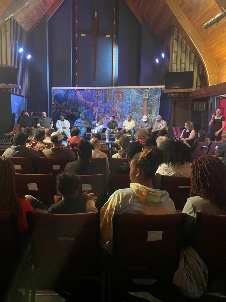 Audience seated in a church-like hall facing a colorful panel on stage with performers