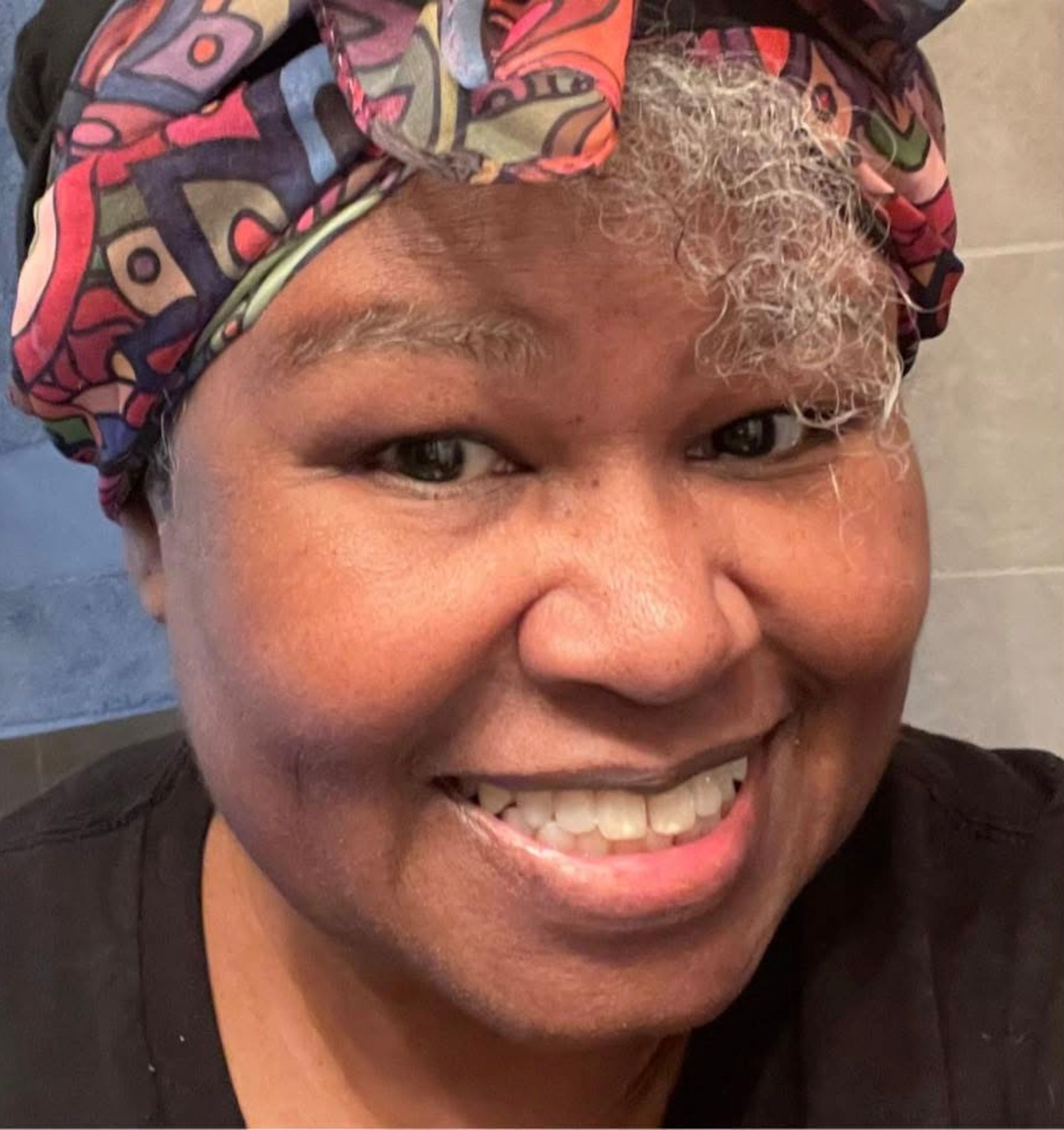 Smiling person in a colorful patterned headwrap and black top, posing indoors against a tiled wall.