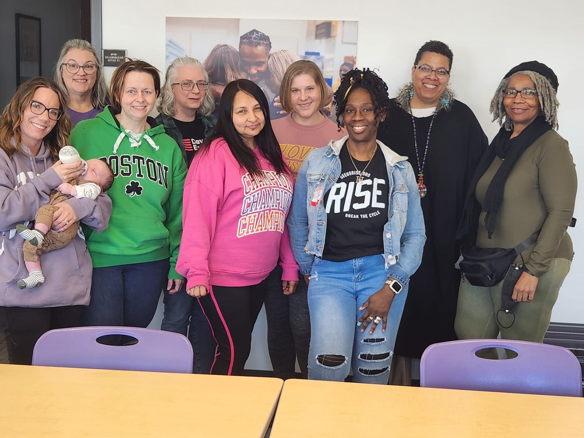 Group of people posing indoors behind a table, smiling in a classroom or meeting room.