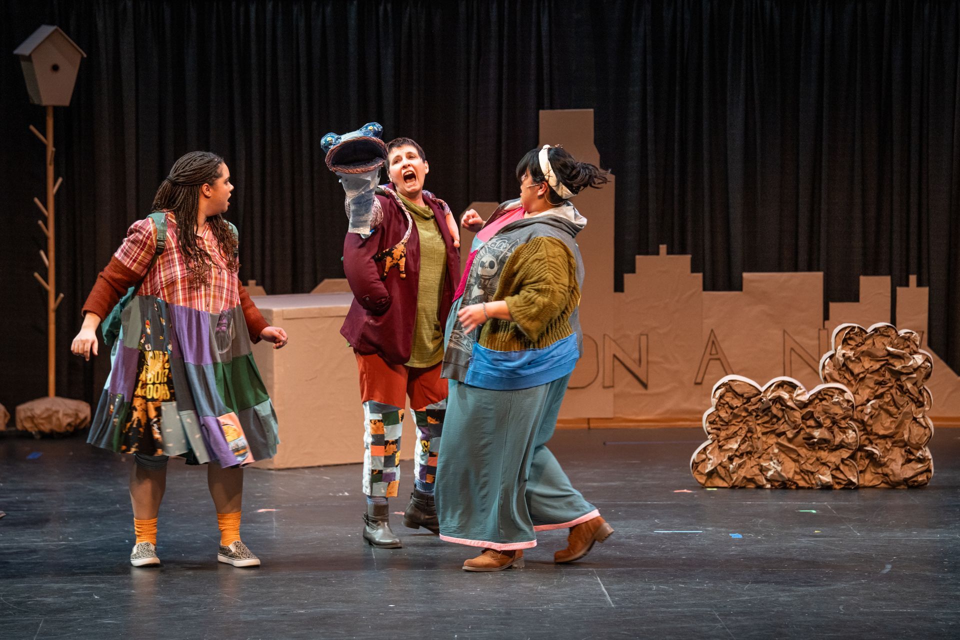 Four actors in colorful costumes on a stage, gathered in conversation before a cardboard city backdrop.