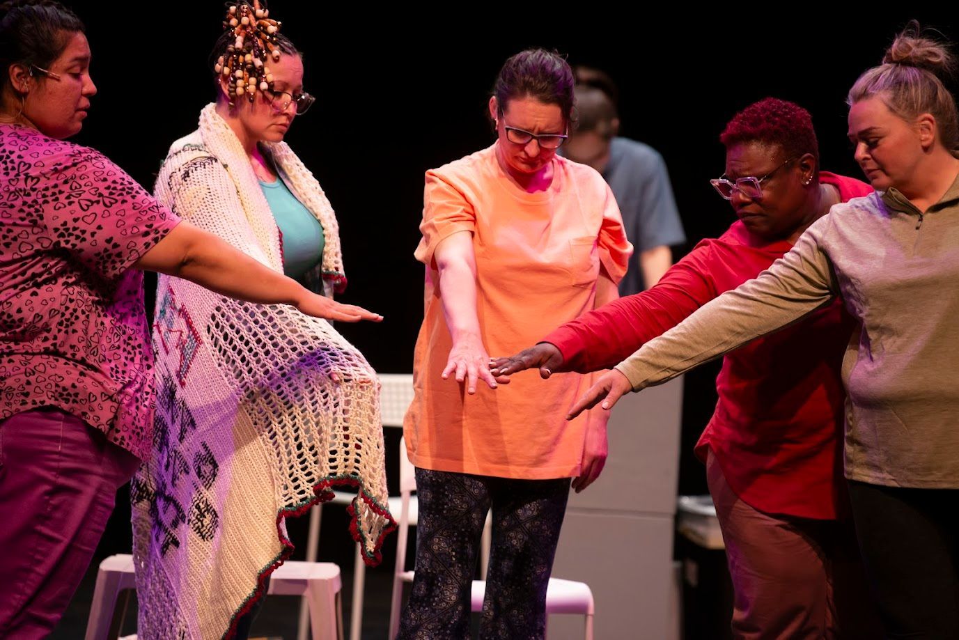 Five people stand onstage in a circle, extending their arms inward in a group gesture.