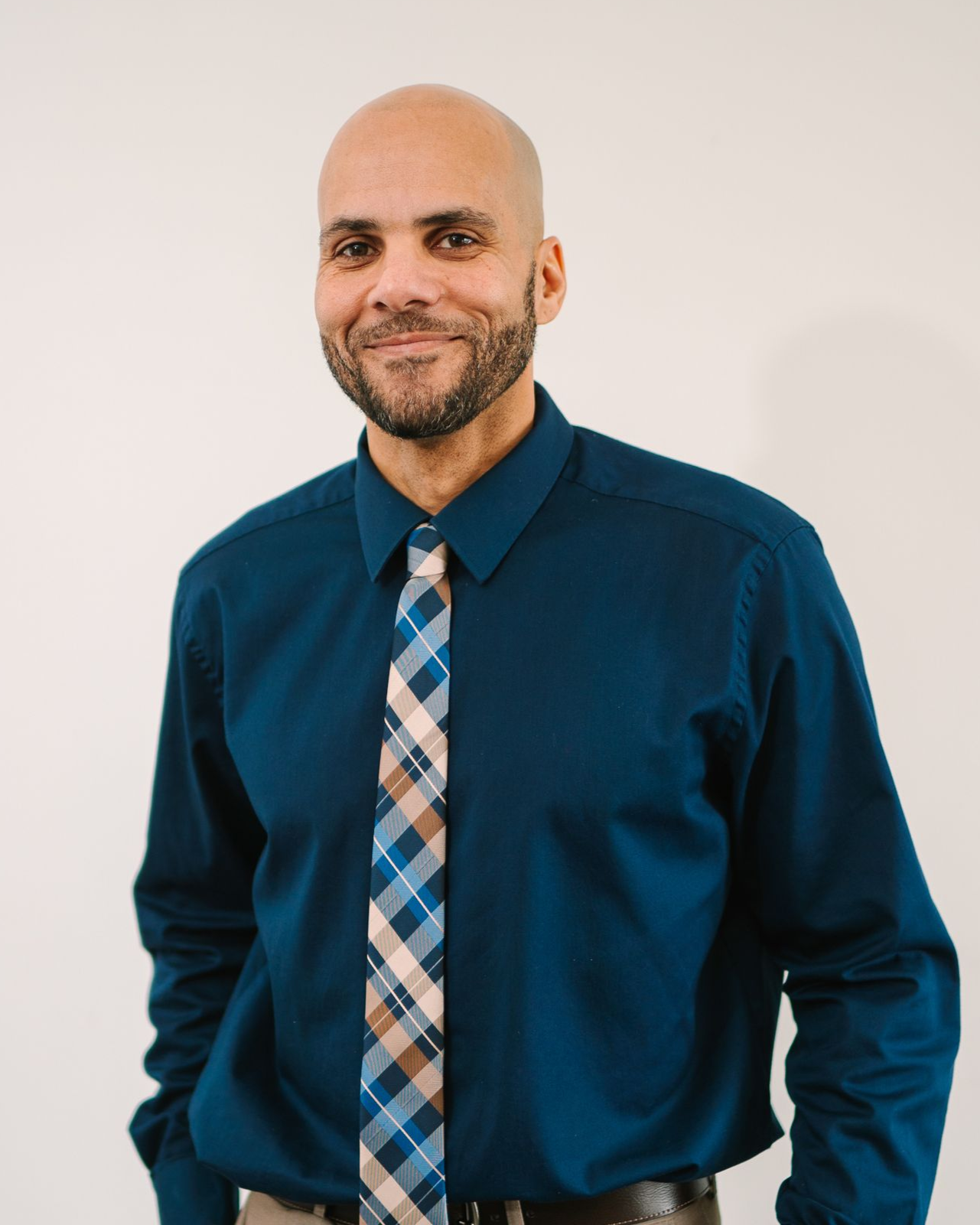 Smiling bald man in a blue shirt and plaid tie, standing against a plain light background