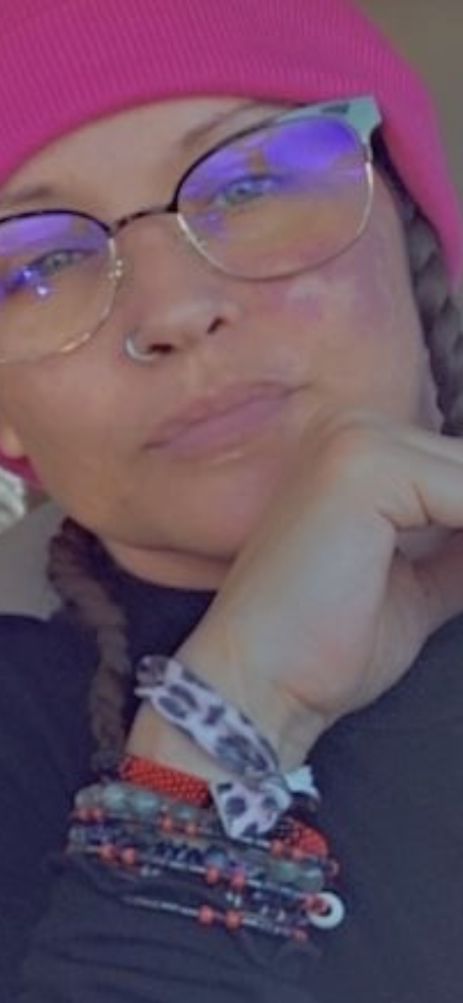 Close-up of a person wearing a pink beanie, glasses, and beaded bracelets, resting their chin on their hand.