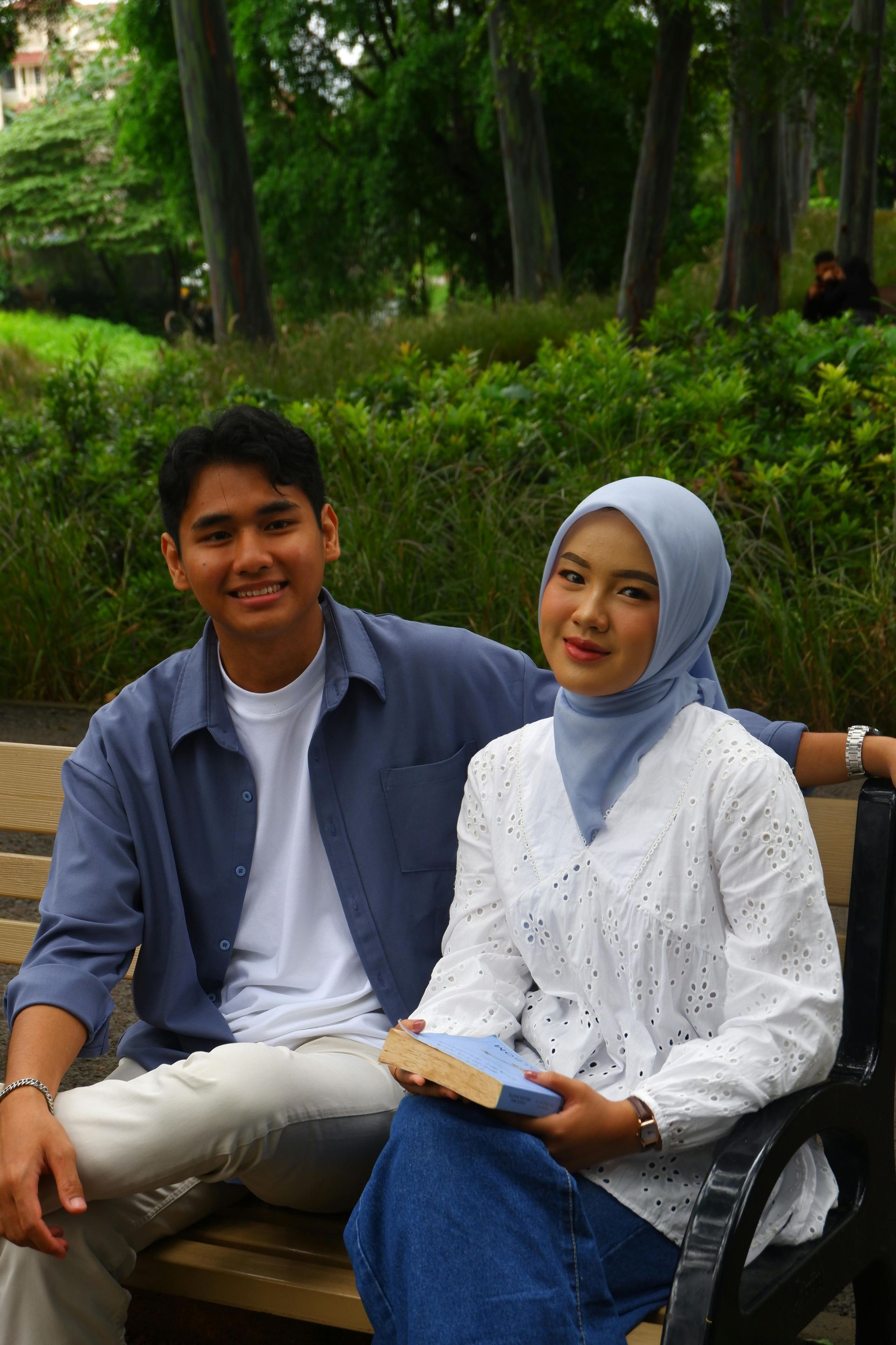 Couple sitting on a park bench. Man in blue shirt, woman in white top and hijab, holding a book.