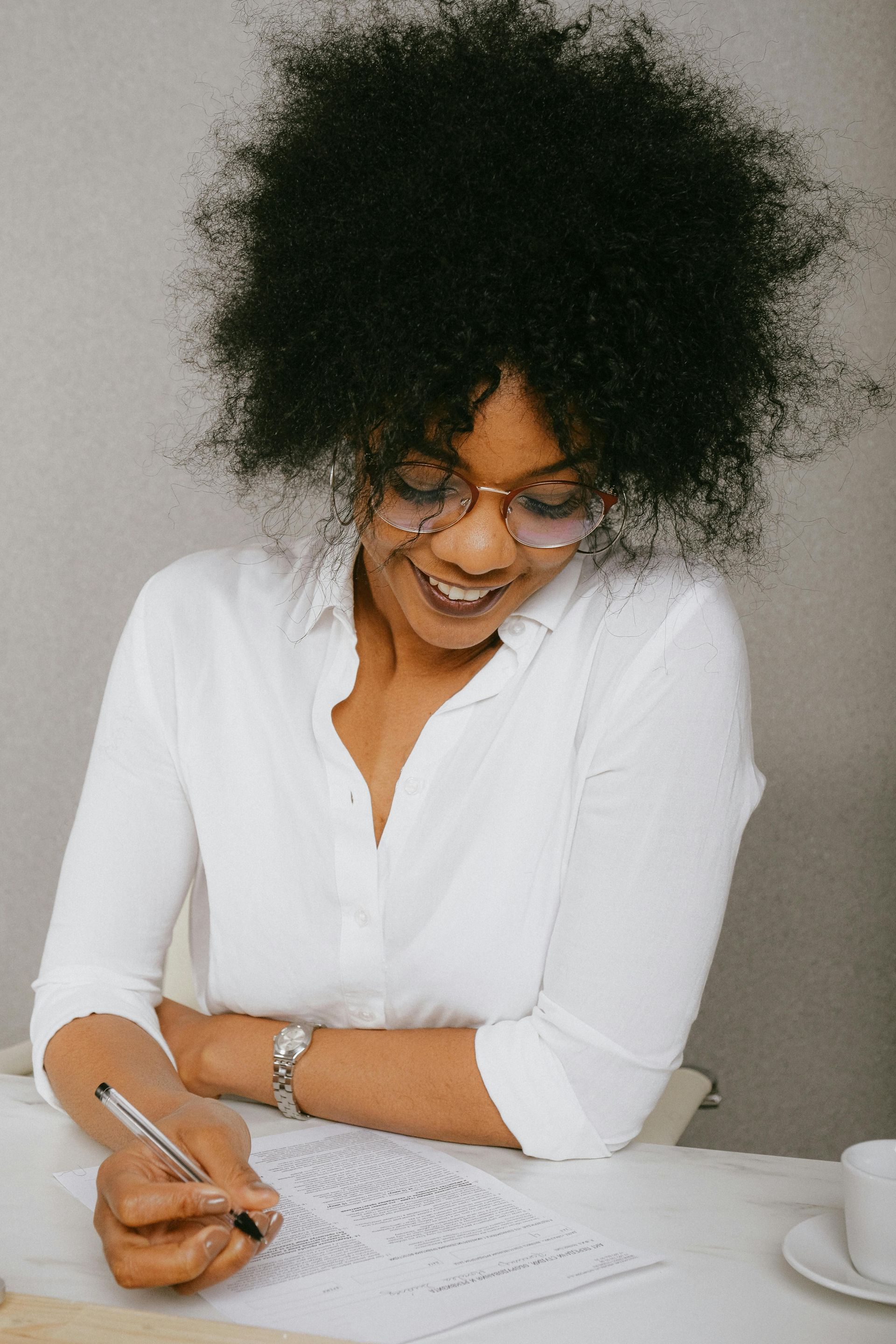 Woman with curly hair, wearing glasses and a white shirt, smiles while writing at a table.
