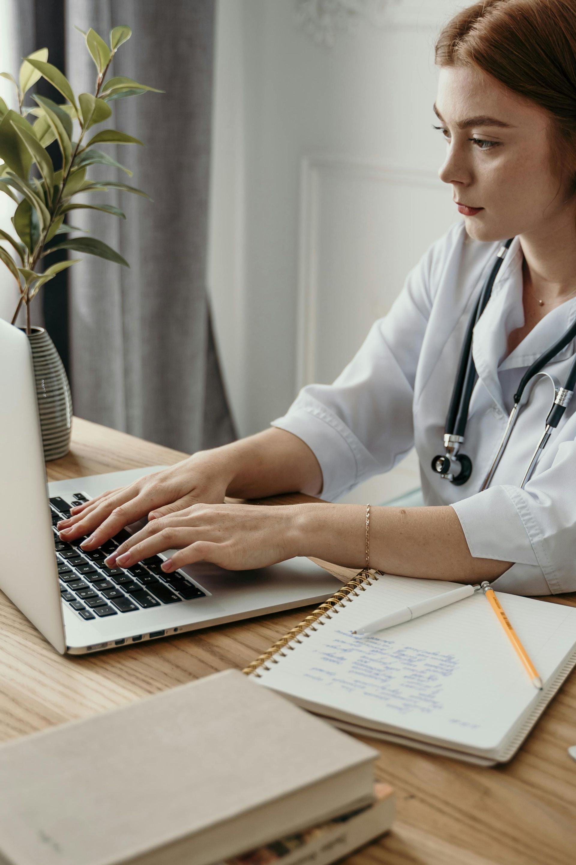 Person in a white coat with a stethoscope typing on a laptop at a desk with a notebook and plant.