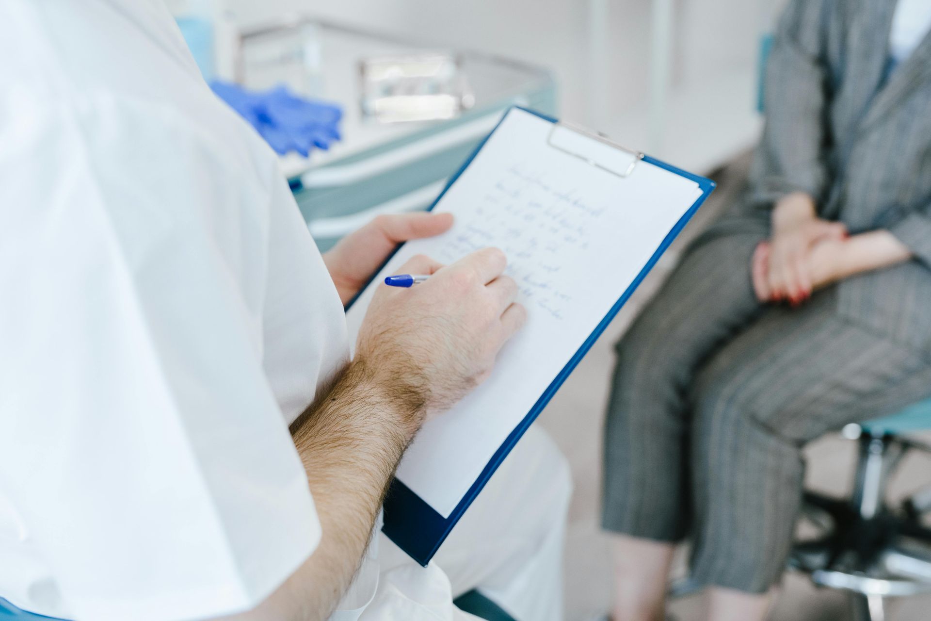 Doctor in white coat taking notes on a clipboard, interviewing a patient seated in a chair.