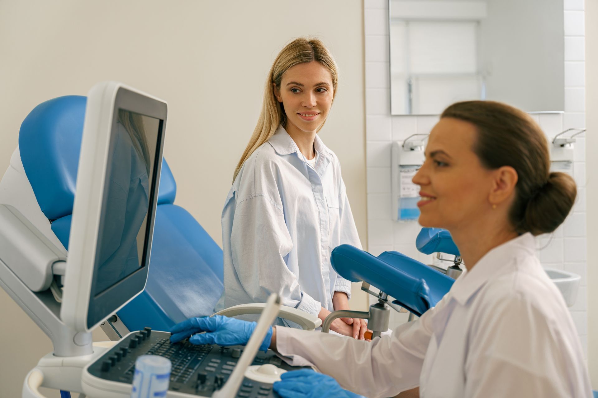 Woman receiving ultrasound in doctor's office. Doctor operates machine, patient smiles. Blue examination chair visible.