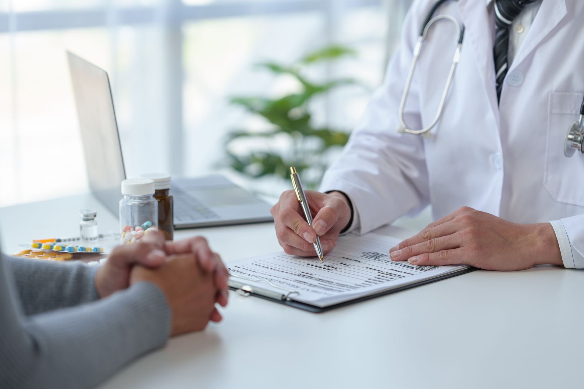 Doctor writing on a clipboard across from a patient; pills, bottles, and laptop on desk.