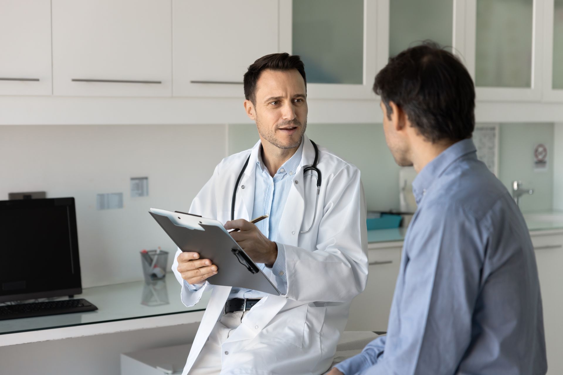 Doctor in white coat consults with patient in a light blue shirt in a medical office, holding a clipboard.