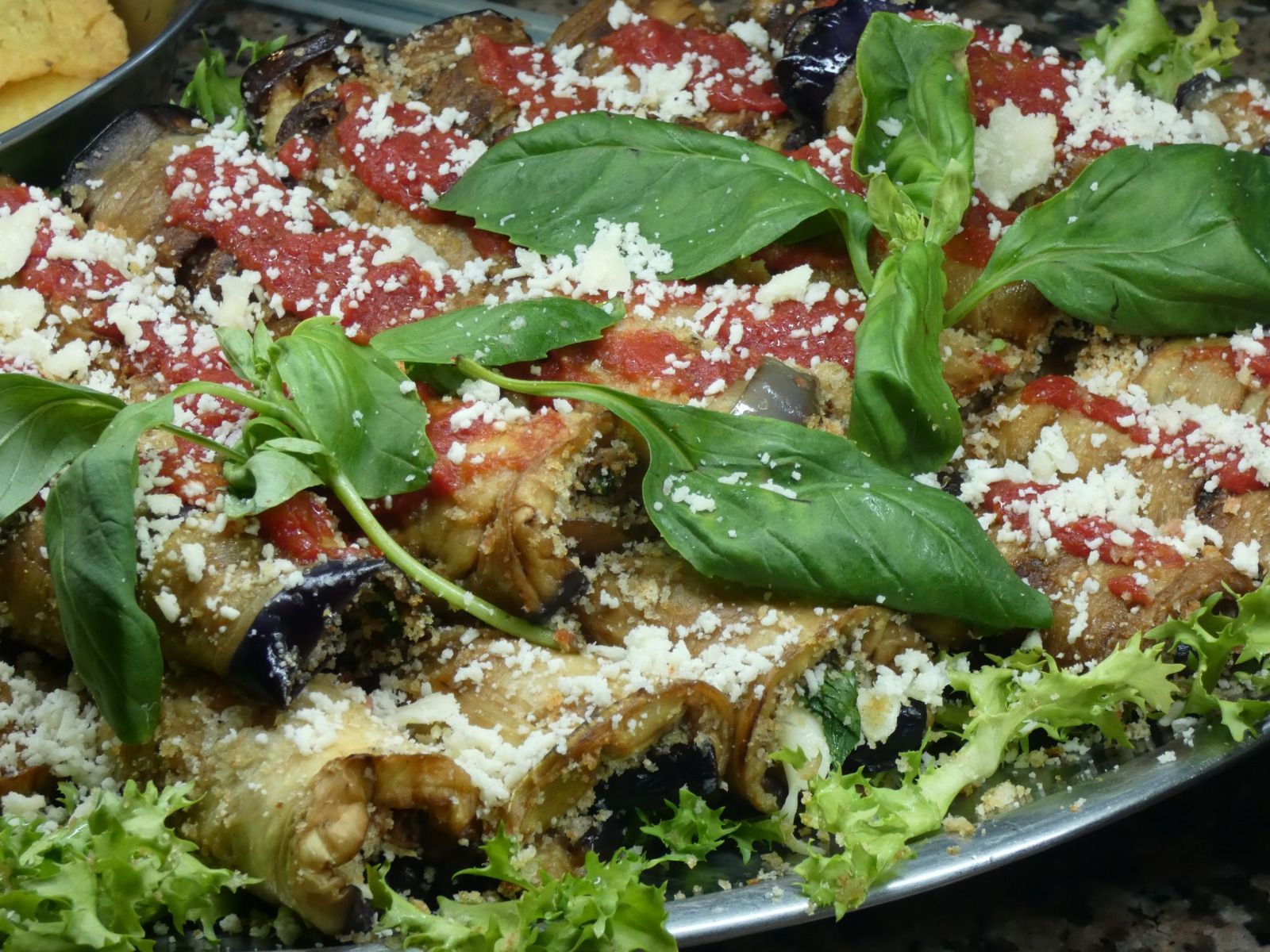 Piatto di involtini di melanzane conditi con salsa di pomodoro, formaggio grattugiato e foglie di basilico fresco su un letto di verdure.