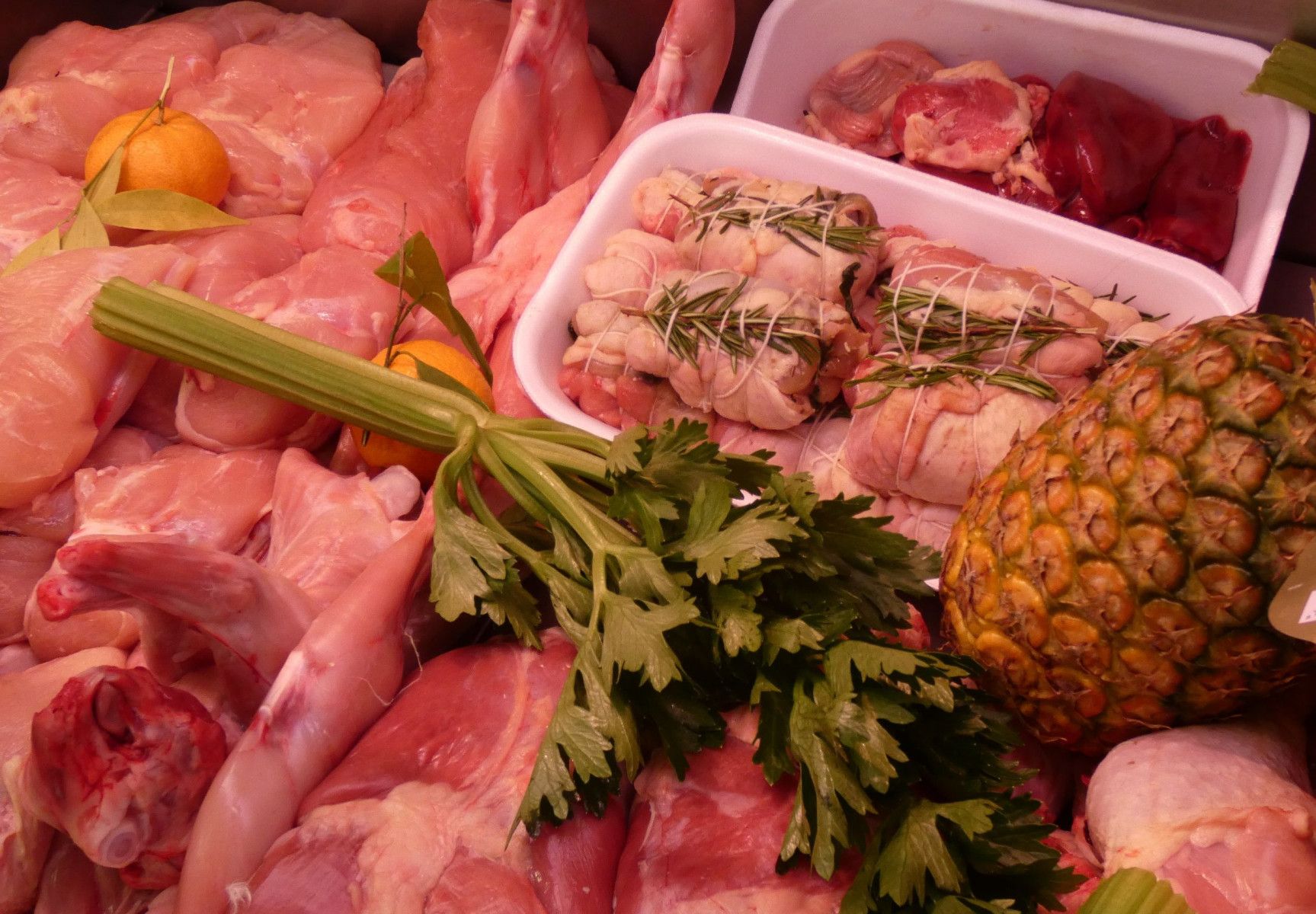 Pezzi di pollo crudo, involtini di carne alle erbe e un ananas sono disposti in una vetrina refrigerata.