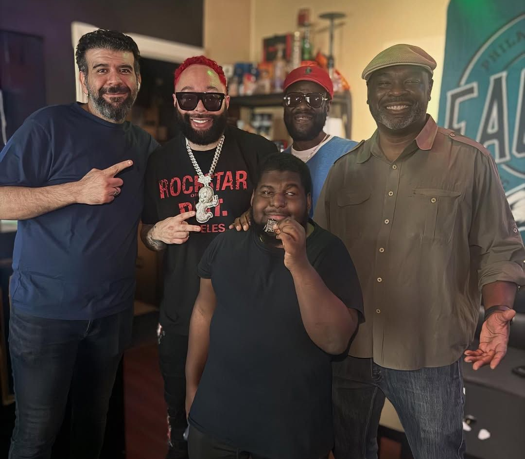 Five people posing together indoors, smiling. One points, one holds a microphone, and another wears a hat.