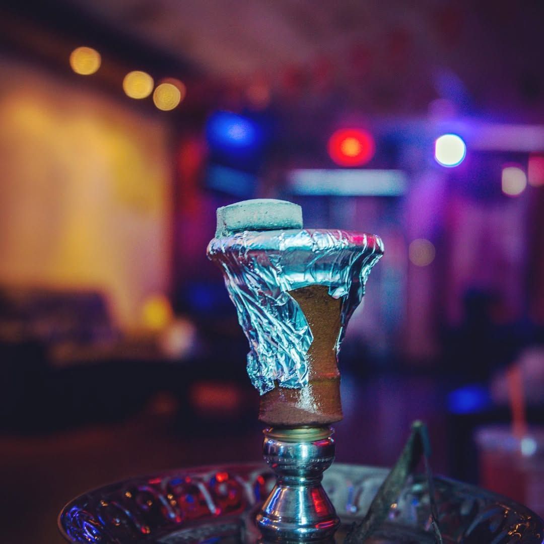Close-up of a hookah with foil-covered bowl and lit charcoal in a dimly lit, colorful lounge.