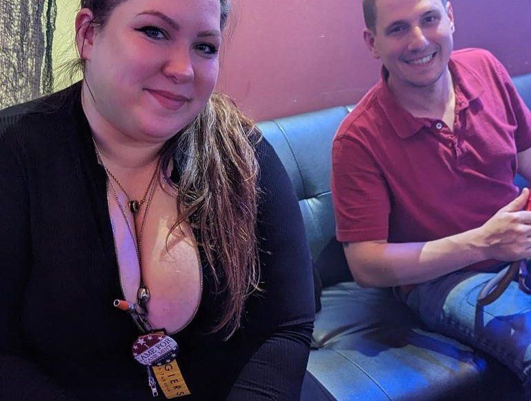 Woman in black top with low neckline, man in red shirt on a black couch, both smiling. Dark setting.