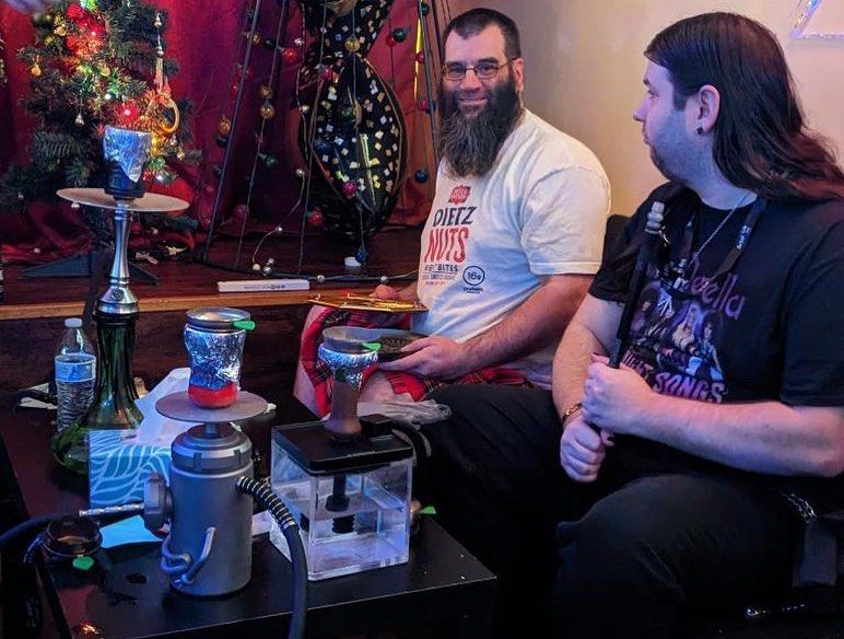 Two men sit, smoking from hookahs, in front of a Christmas tree. One smiles.