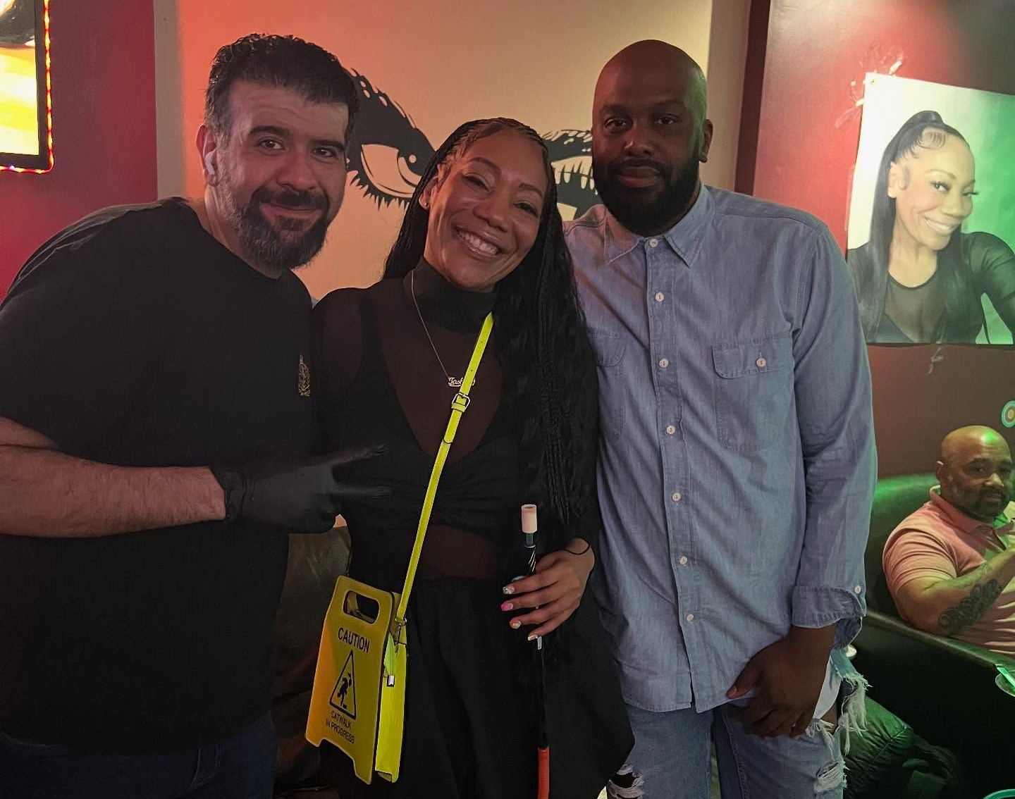 Three people smiling inside a lounge; two men and a woman. The woman holds a cane and yellow bag. One man wears black gloves.
