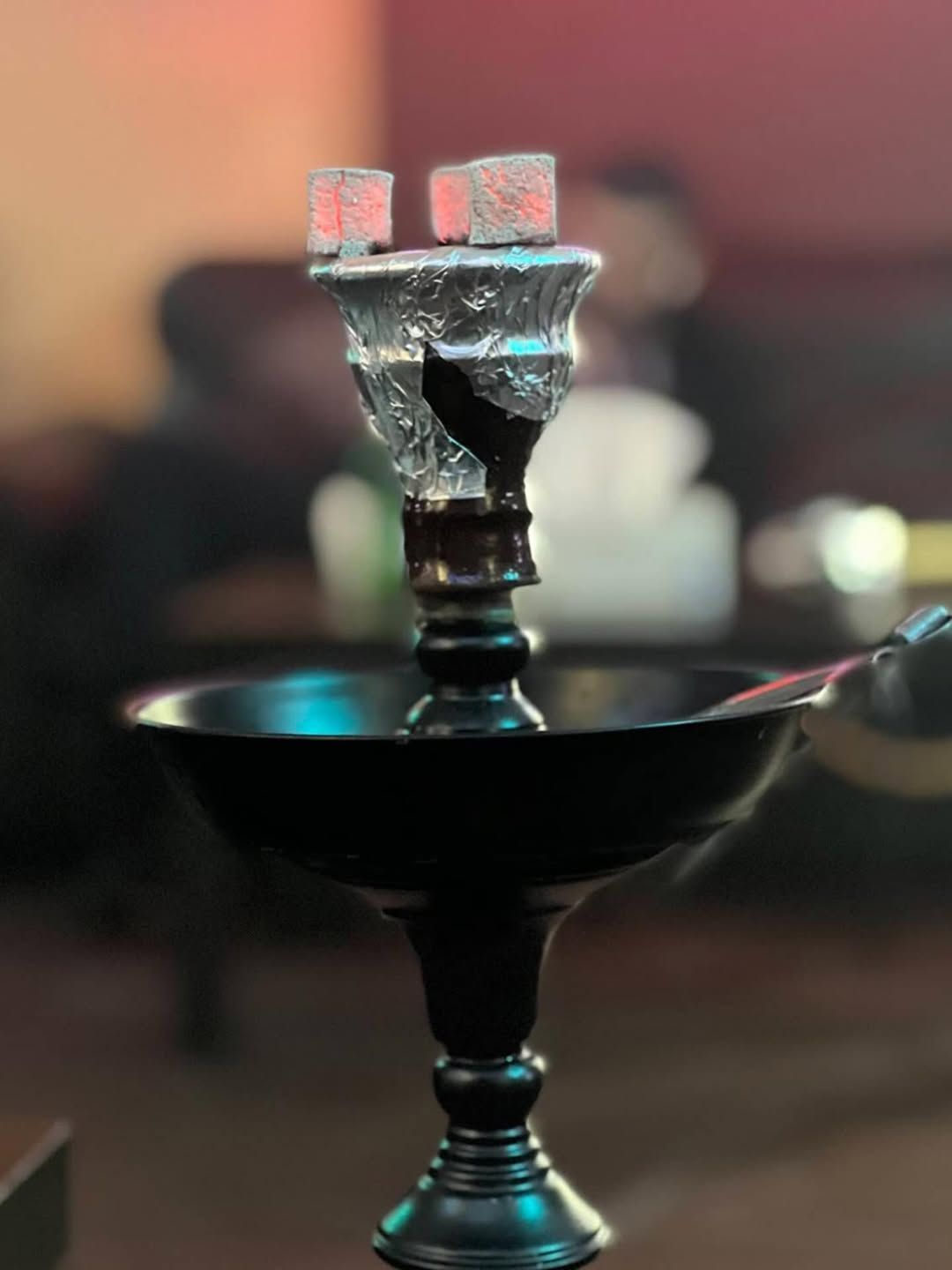 A hookah with glowing coals, set indoors. Black base, blurred background.