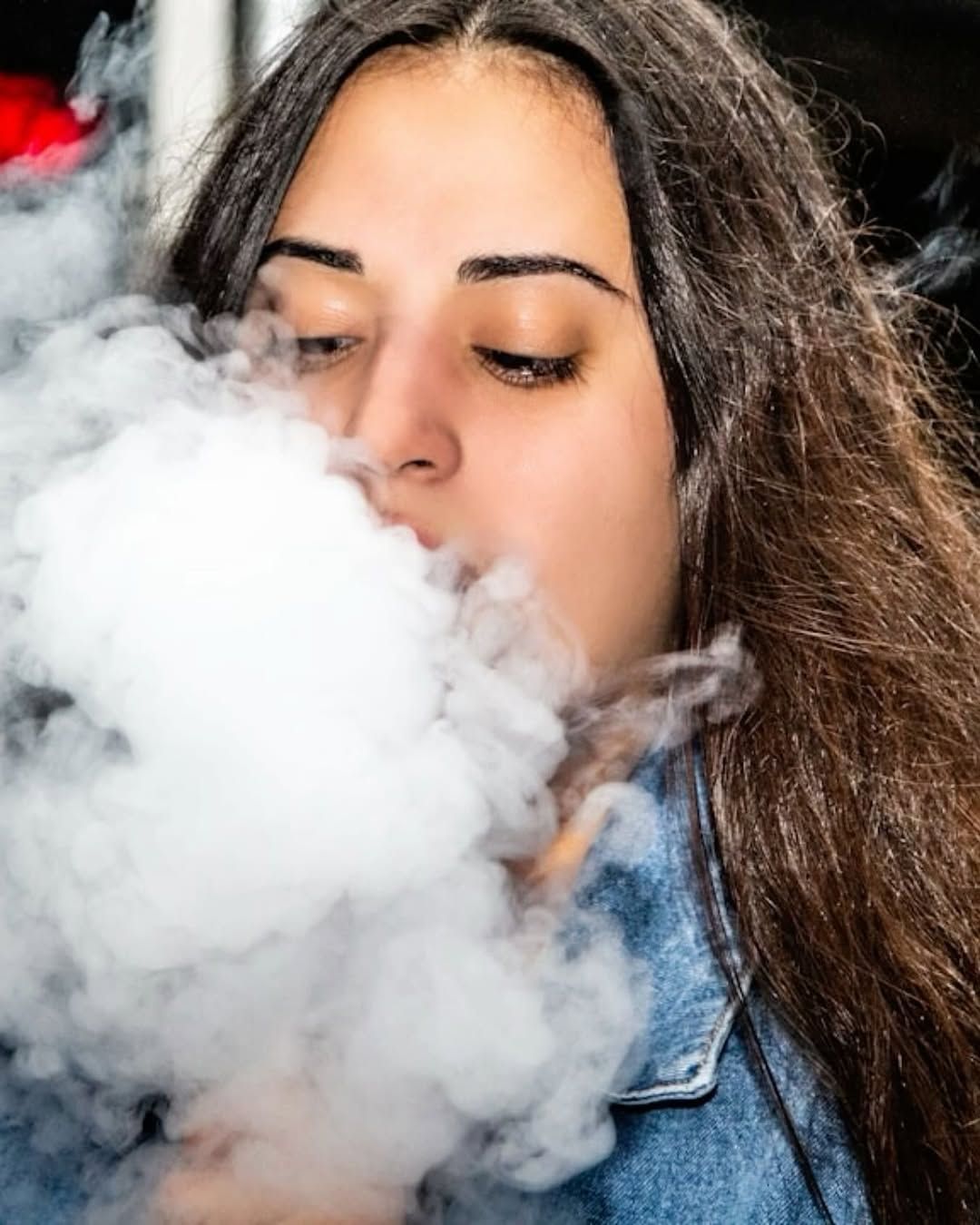 Woman exhaling a large cloud of white vapor, wearing a denim jacket.