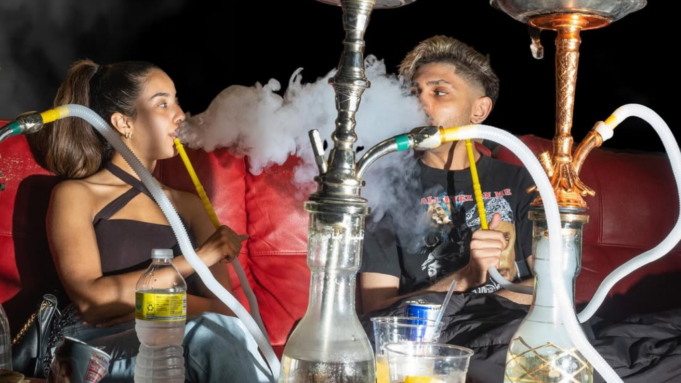 Two people smoking hookah, exhaling large plumes of smoke. Seated on red sofa, dark background.