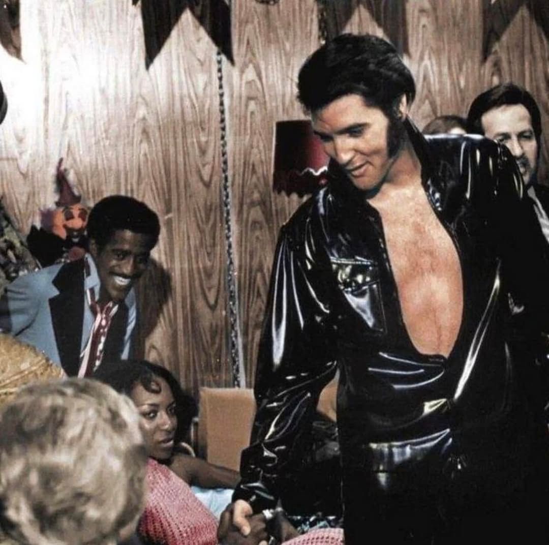 Elvis Presley im schwarzen Vinyl-Jumpsuit, lächelnd, umgeben von Menschen, in einem Innenraum.