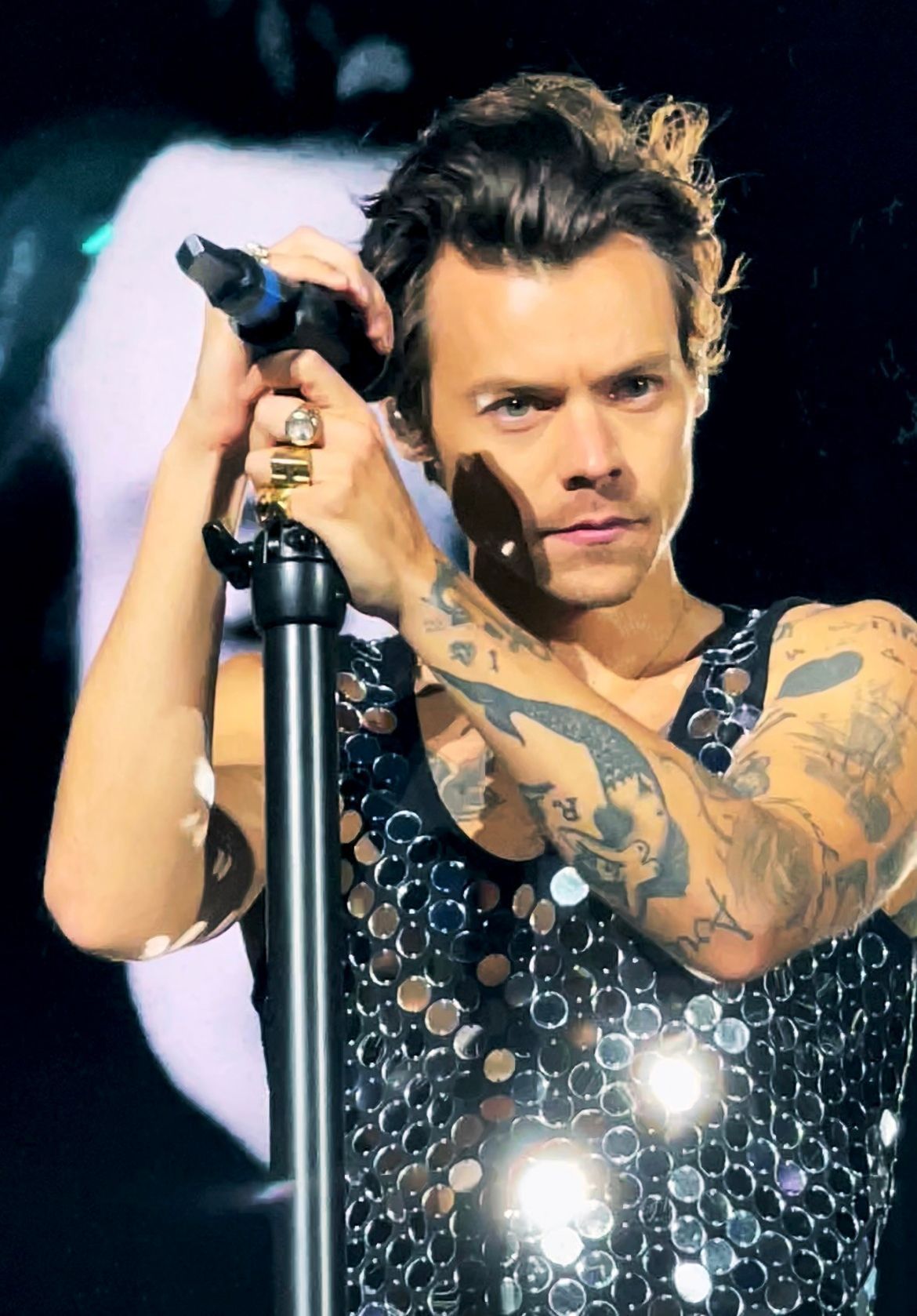Singer holding a microphone onstage, wearing a black sequined top with tattooed arms visible.