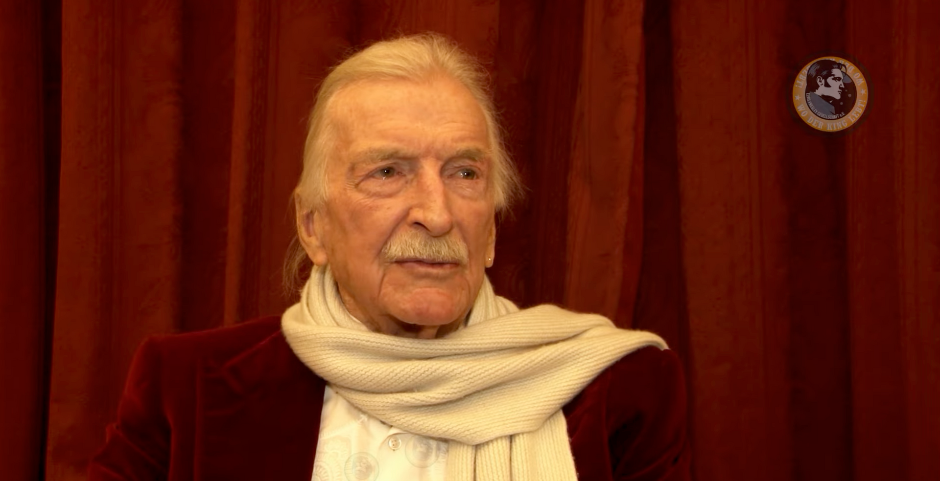 James Last trägt eine rote Jacke und sitzt vor roten Vorhängen.