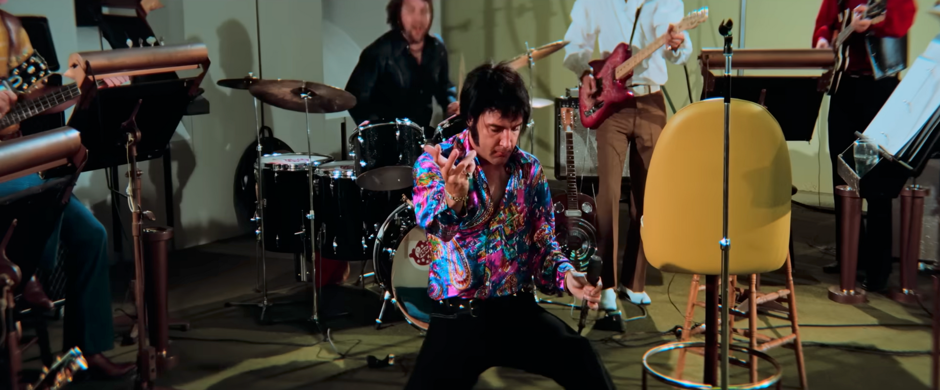 Elvis Presley tritt mit seiner Band auf. Er trägt ein buntes Hemd und eine schwarze Hose und gestikuliert mit den Händen.