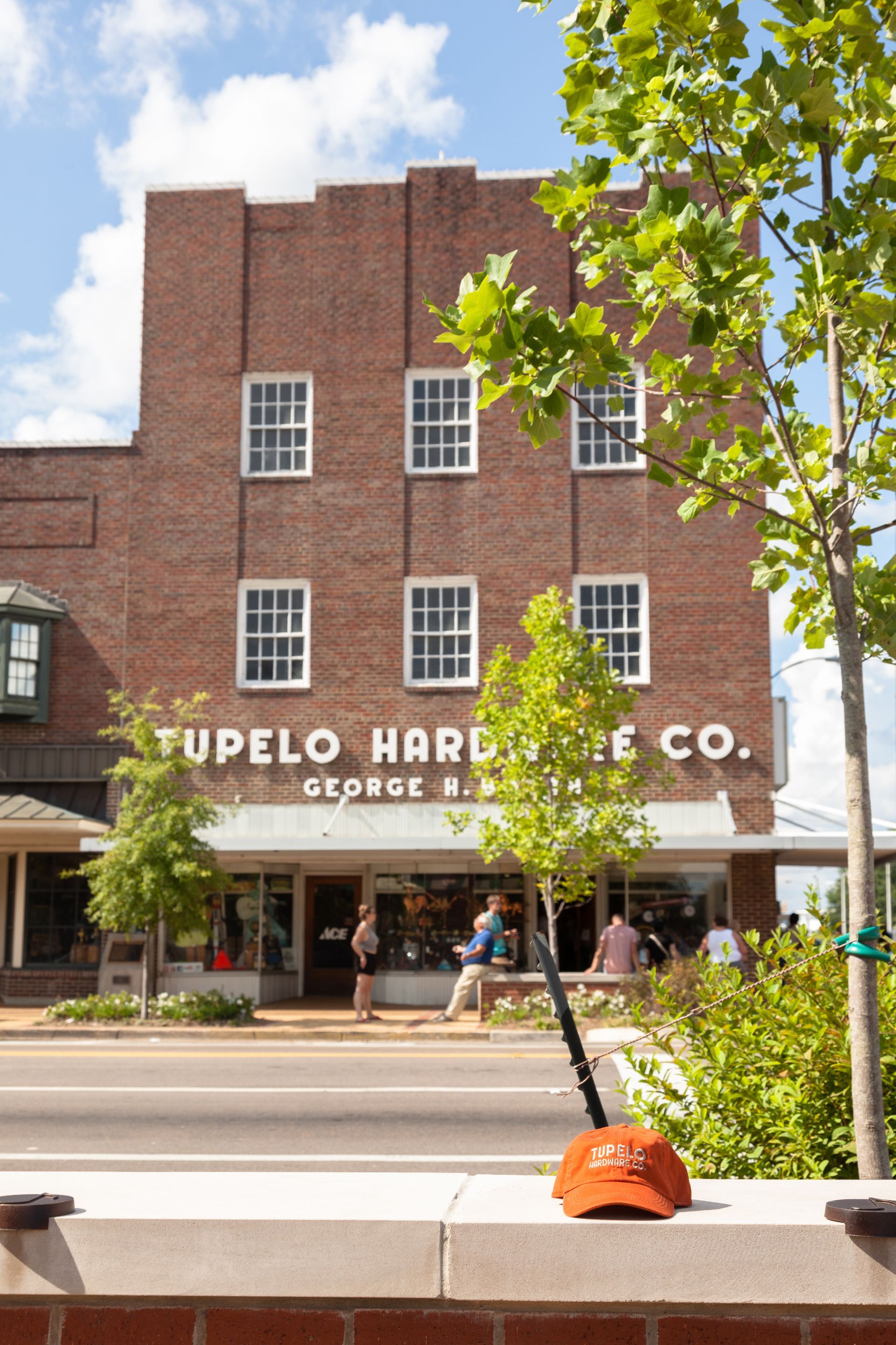 Das Backsteingebäude der Tupelo Hardware Co. mit weißen Zierelementen, Schaufensterfront und Passanten auf dem Bürgersteig. Auf einer niedrigen Backsteinmauer sitzt eine orangefarbene Abdeckung.