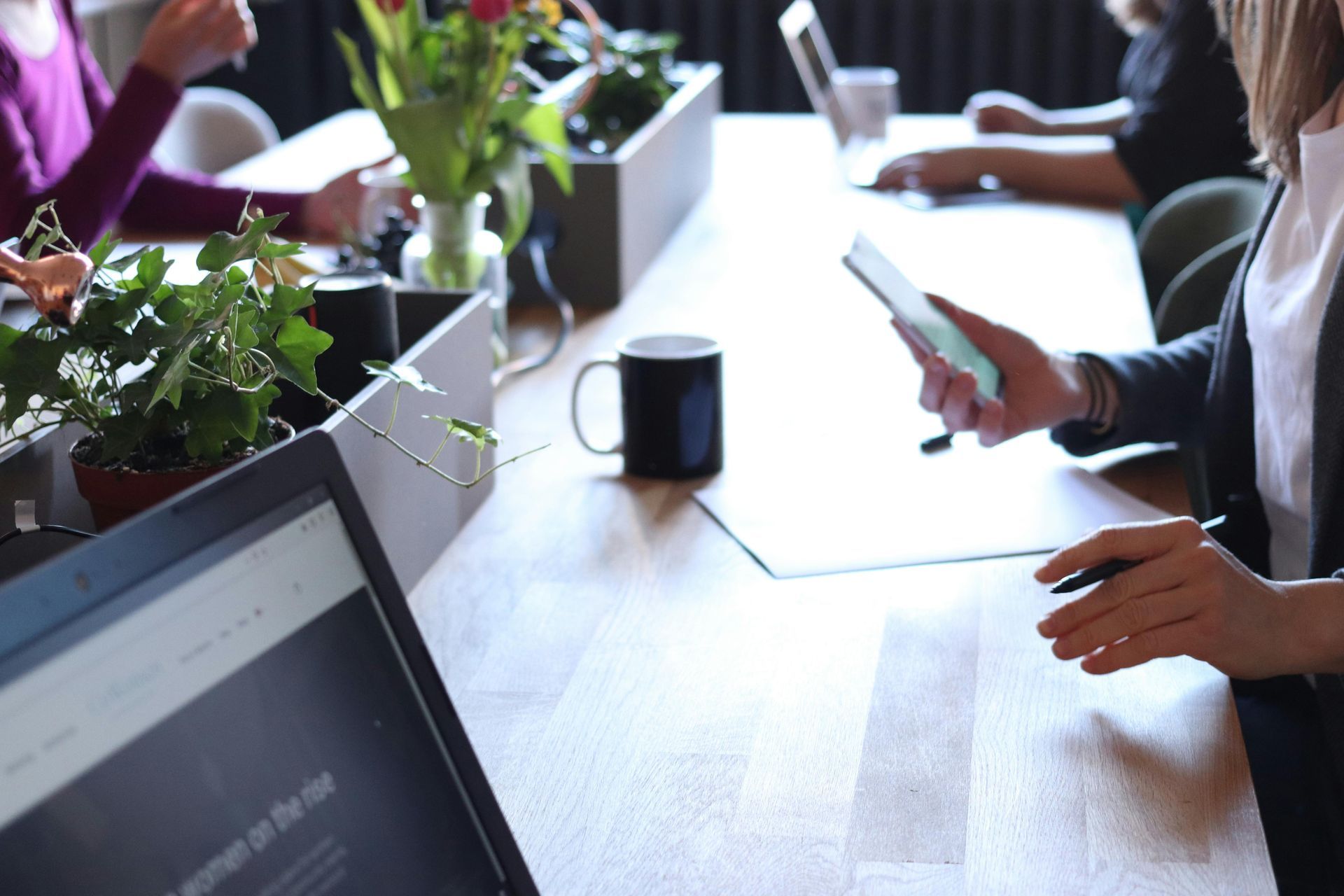 People working at a table with laptops, phones, a coffee mug, and plants.