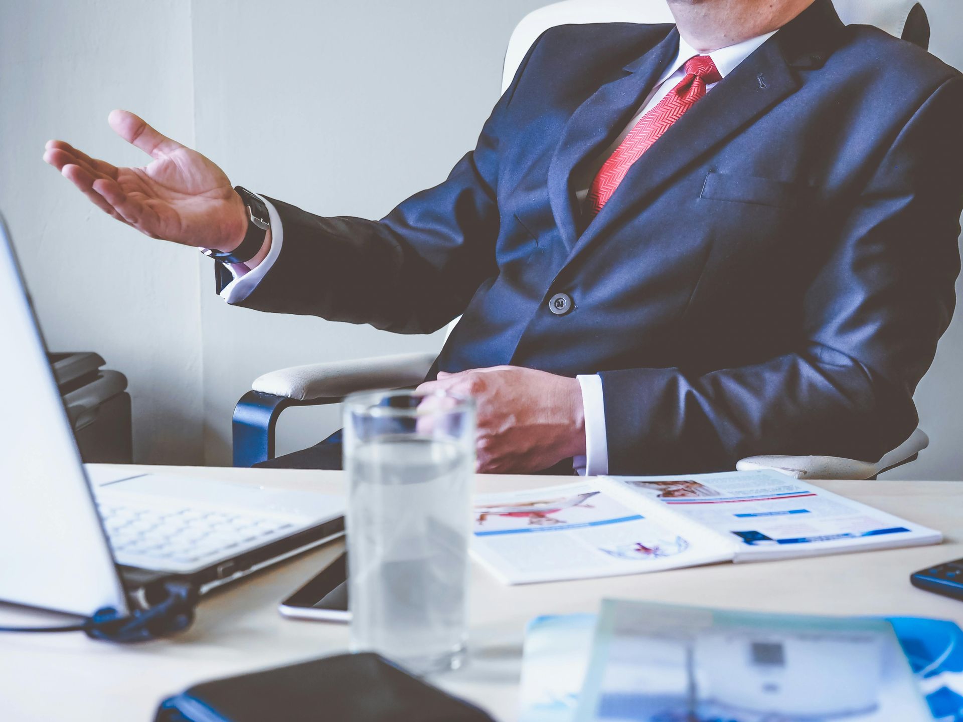 Man in a suit gestures with his hand at a desk with a laptop, reports, and a glass of water.