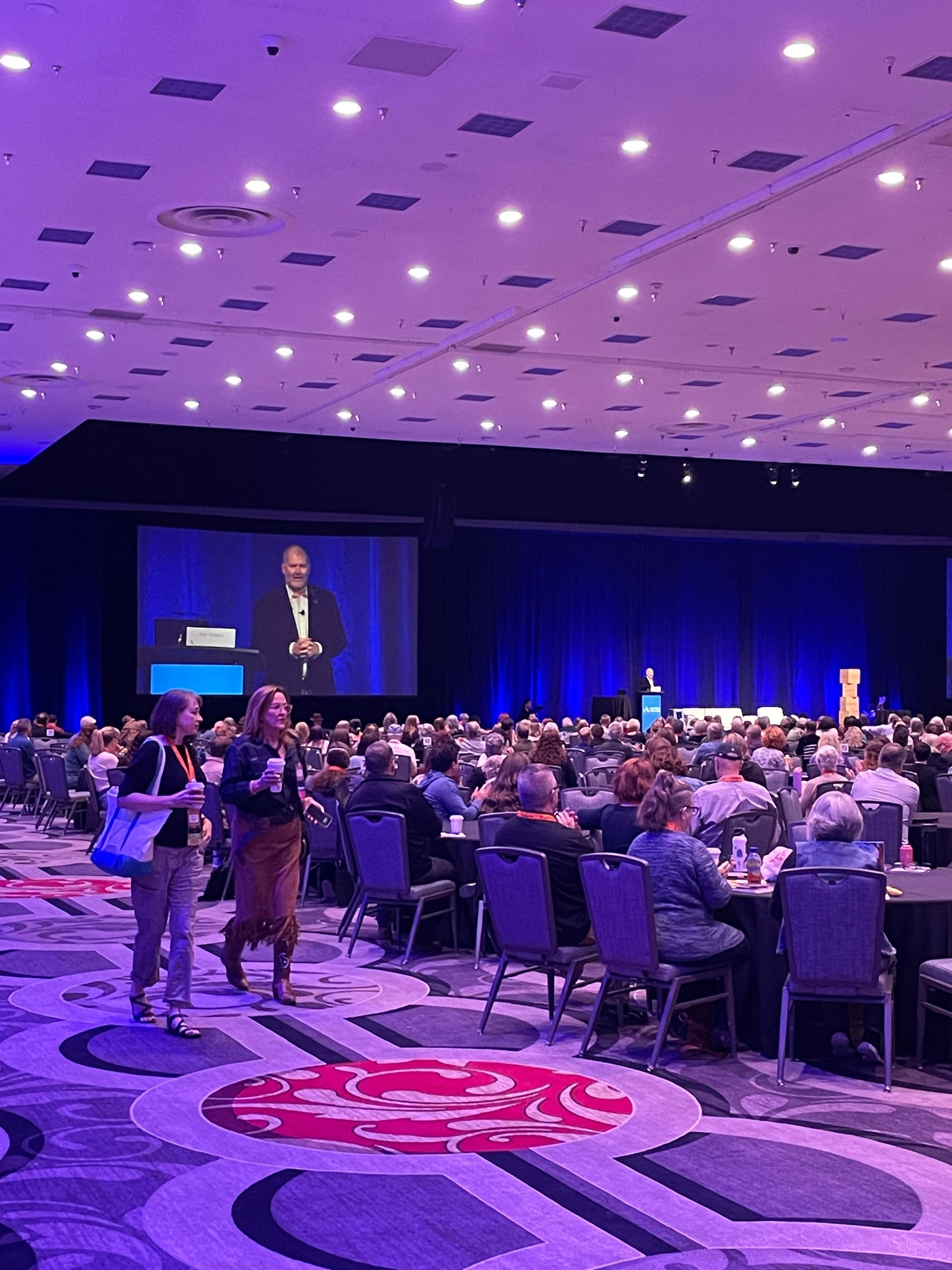 Large conference hall filled with people. A speaker on stage; audience seated at tables. Purple lighting.