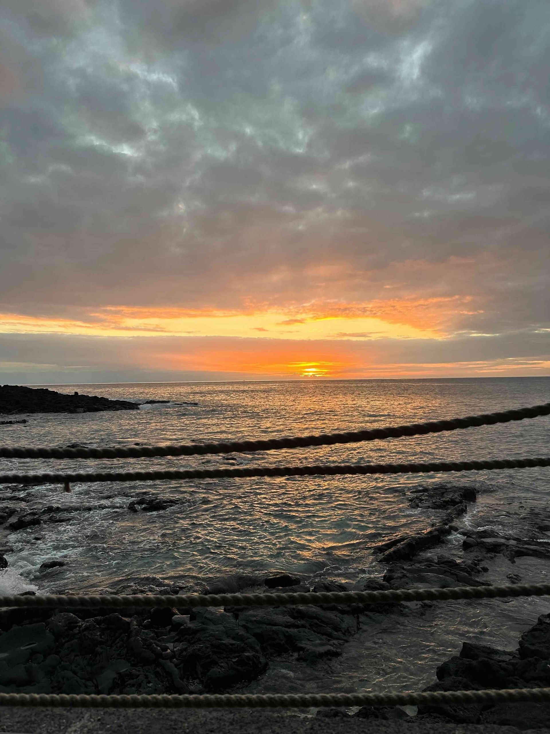 Sunset over ocean with orange and gray sky; rope fence in foreground.