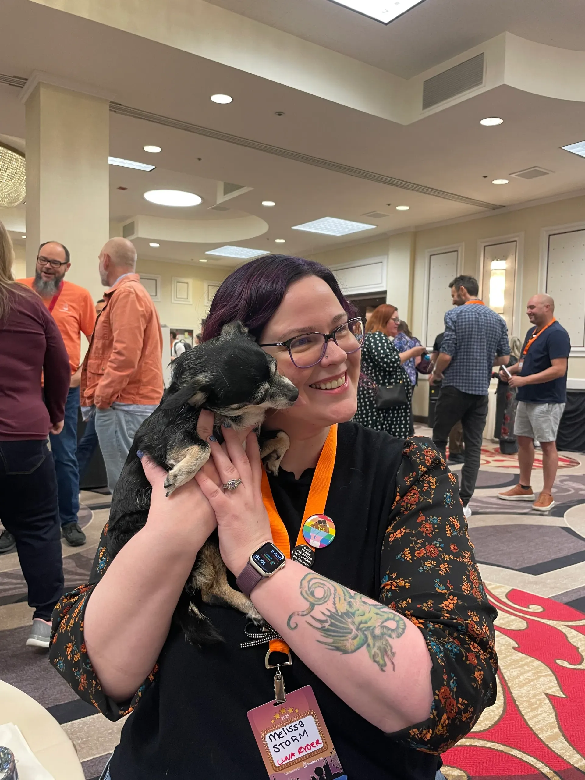 Woman with purple hair smiles, holding a small black and tan dog in a brightly lit room with people mingling.