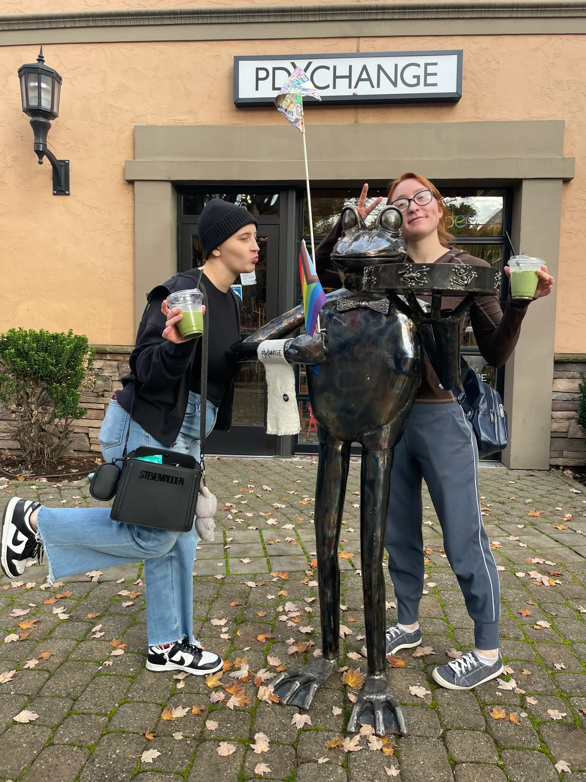 Two people pose with a frog sculpture, holding drinks. Outside a shop named