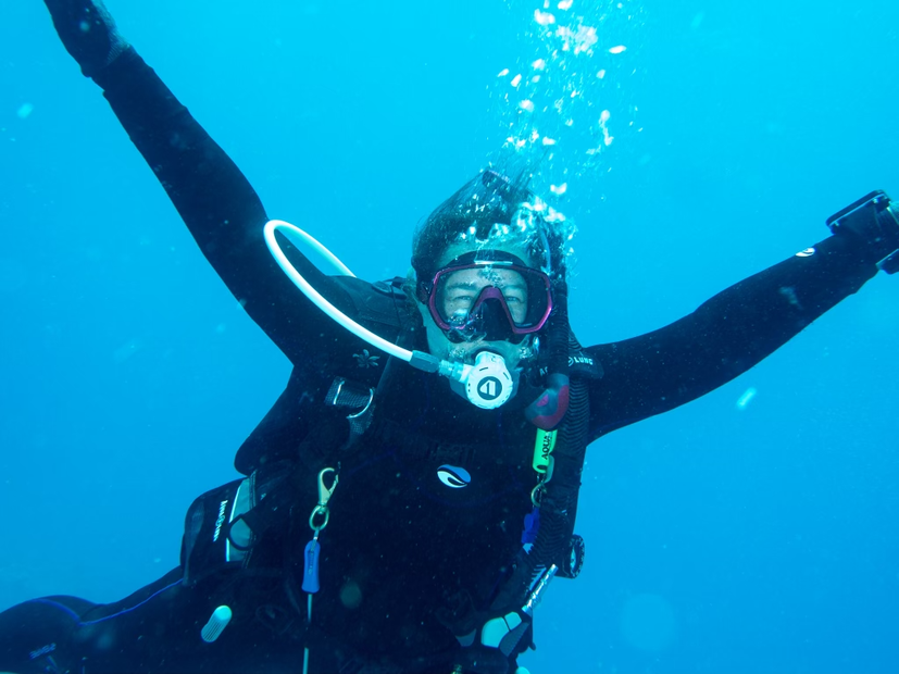 Scuba diver with arms raised, underwater, smiling, wearing black wetsuit and equipment. Blue water background.