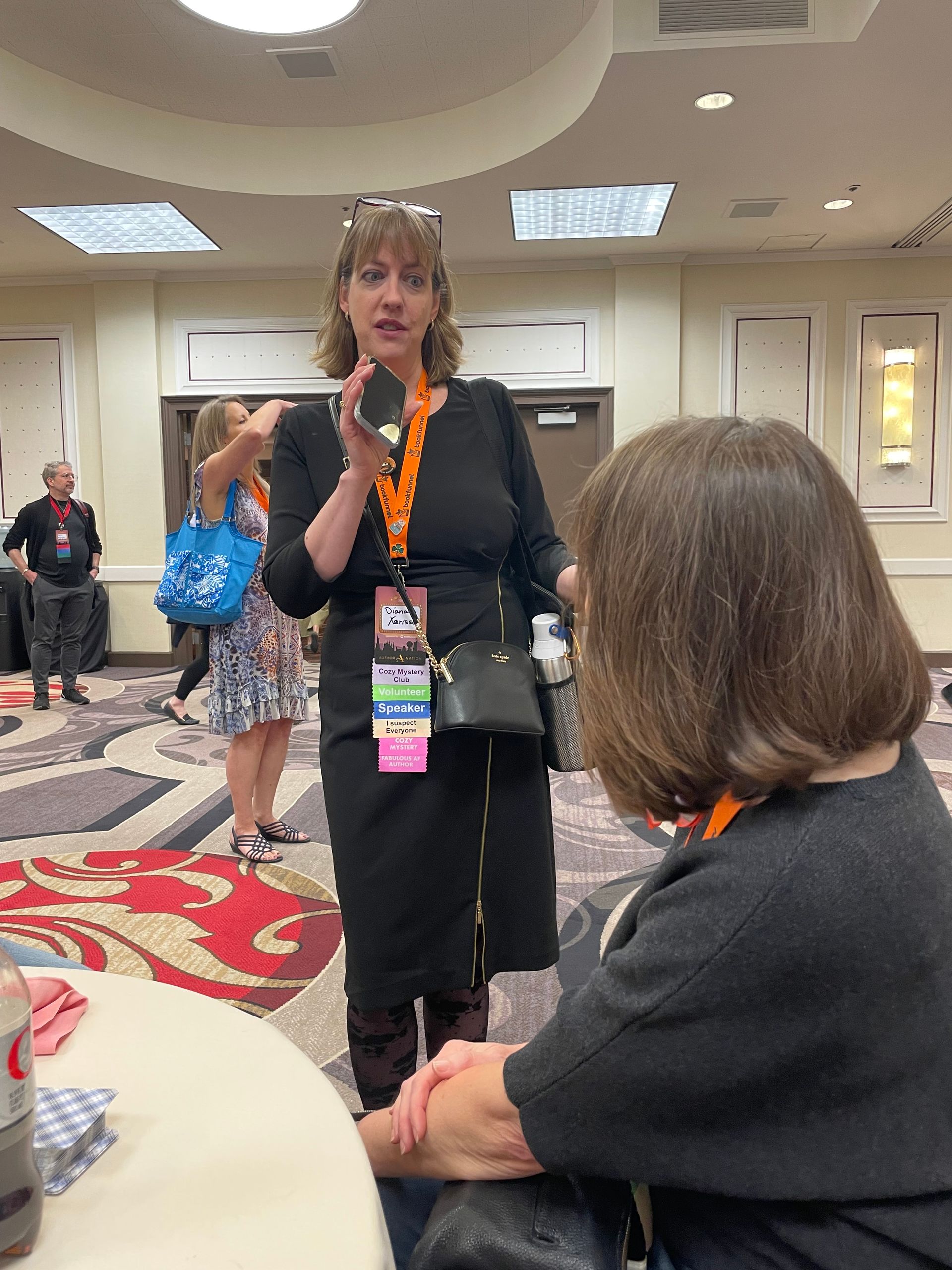 Woman in black dress talking, holding a compact mirror. Sitting person listens. Convention hall setting.